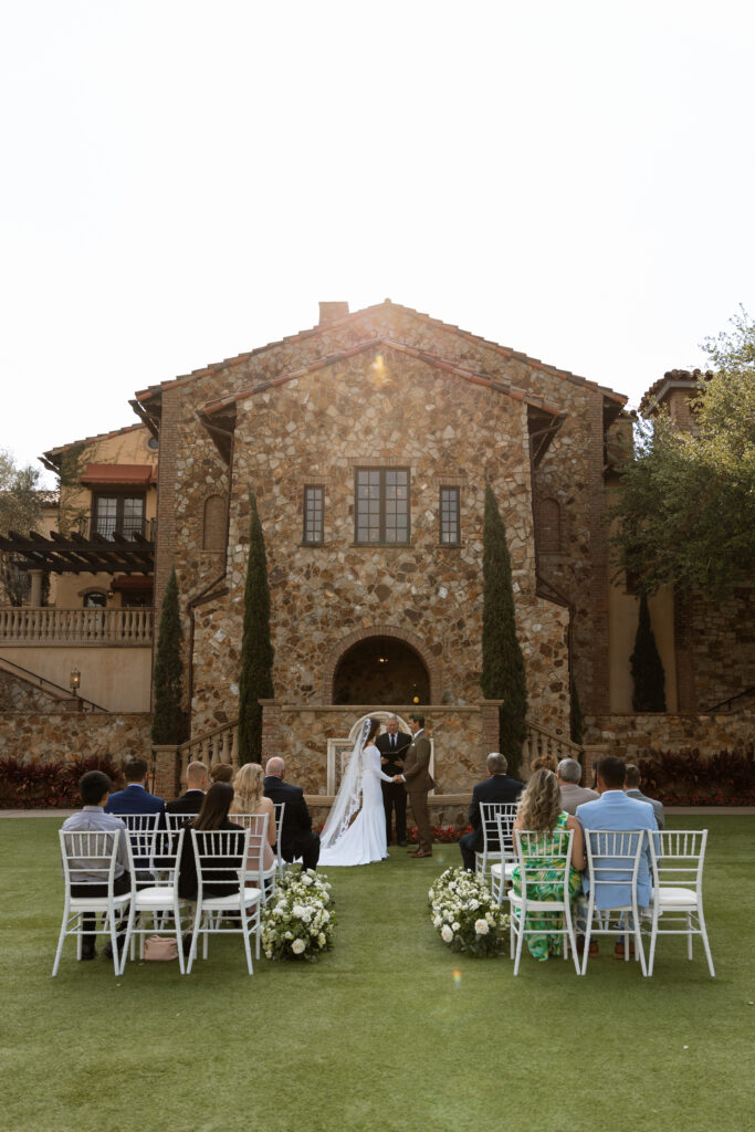 Outdoor wedding ceremony at Bella Collina, photographed by The Deans Photography, with guests in front of the picturesque Tuscan-inspired venue and lush rolling hills.