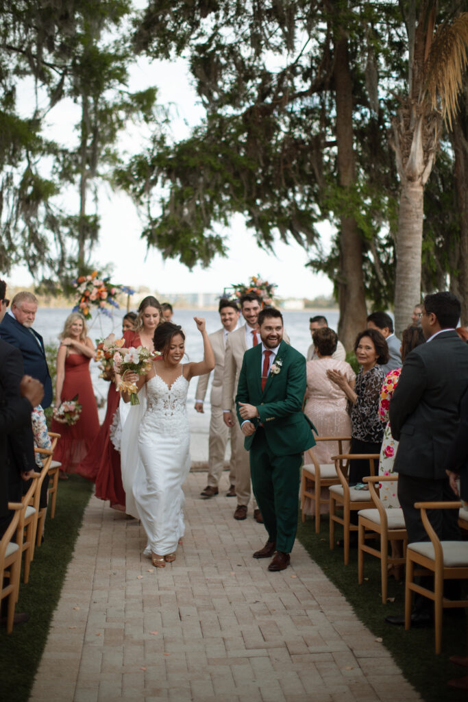 Outdoor wedding ceremony at Paradise Cove, photographed by The Deans Photography, with the couple exchanging vows on the lakeside beach with guests seated on tiki-style chairs.