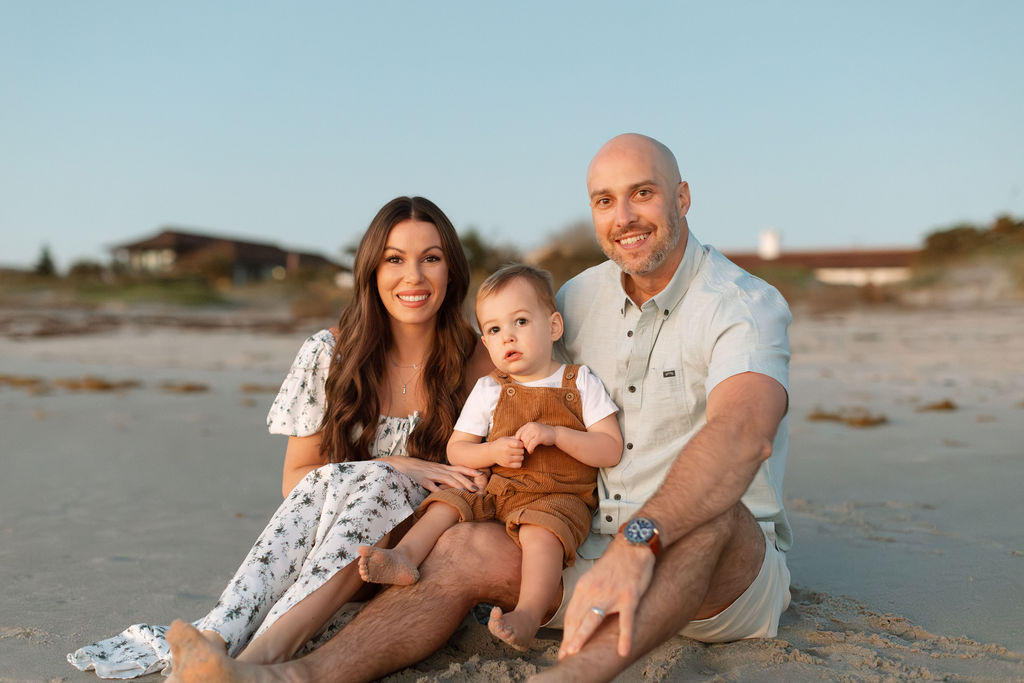 Happy family of three posing together on Cocoa Beach during a sunrise photo session, with colorful sky and soft light.