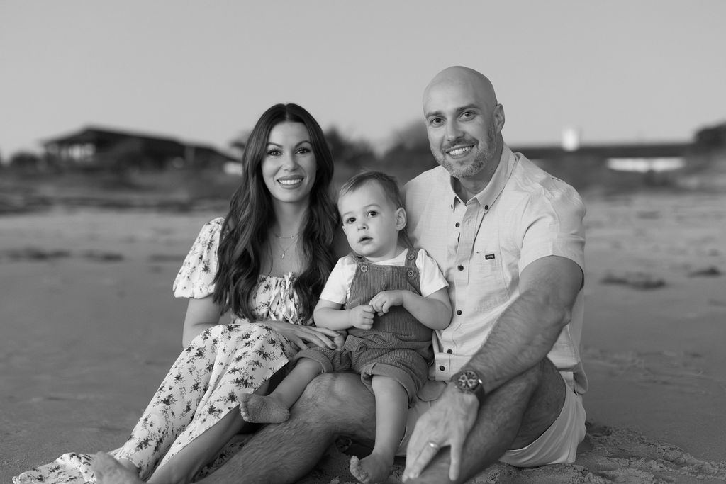 Happy family of three posing together on Cocoa Beach during a sunrise photo session, with colorful sky and soft light.