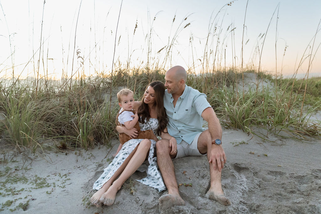 Family of three sitting together on Cocoa Beach during a sunrise photography session, with gentle waves and early morning light.