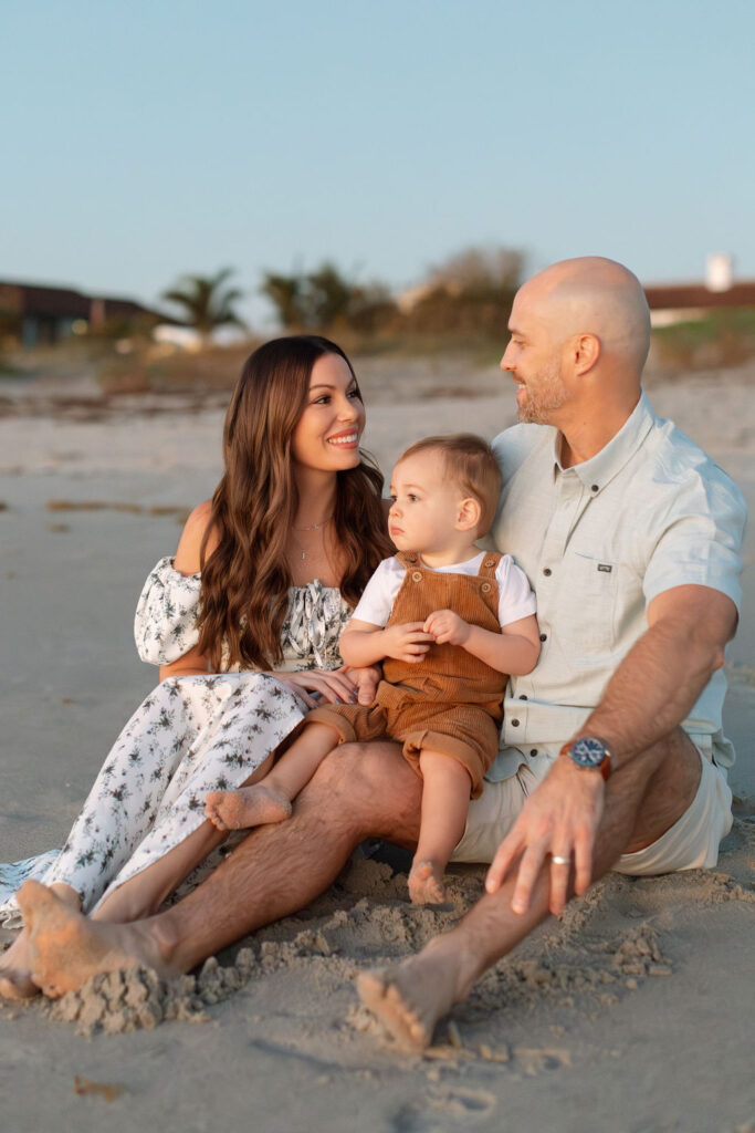 Happy family of three posing together on Cocoa Beach during a sunrise photo session, with colorful sky and soft light.