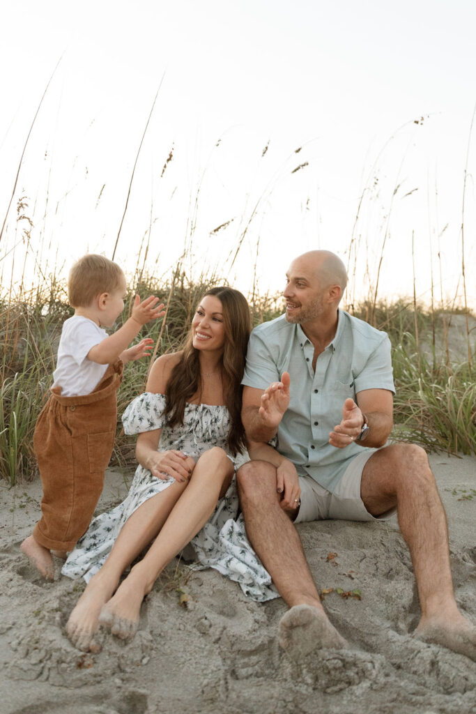 Family of three sitting together on Cocoa Beach during a sunrise photography session, with gentle waves and early morning light.