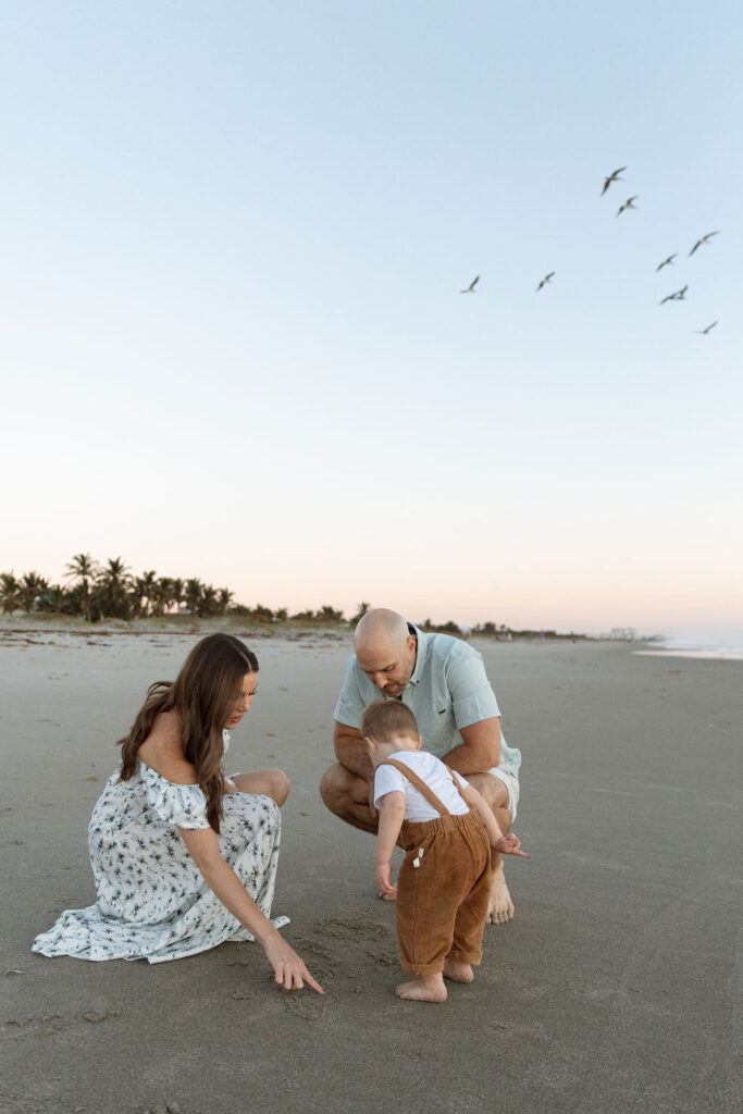 Family of three sitting together on Cocoa Beach during a sunrise photography session, as a flock of birds fly overhead.