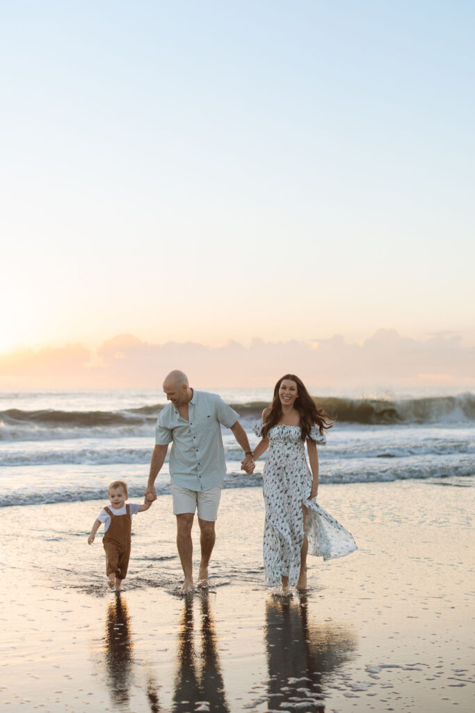 Family of three walking hand-in-hand along Cocoa Beach at sunrise, with soft golden light illuminating their faces.