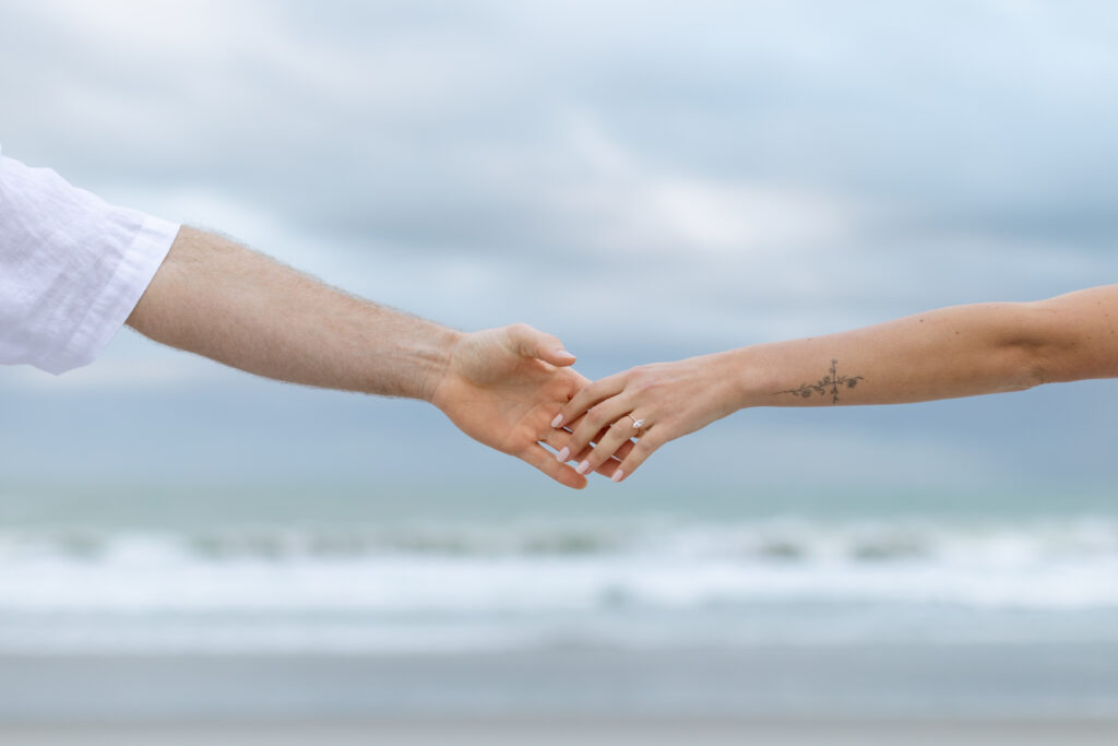 Engaged couple holding hands on Cocoa Beach, with sunset colors in the background, captured by The Deans Photo for a romantic engagement session.