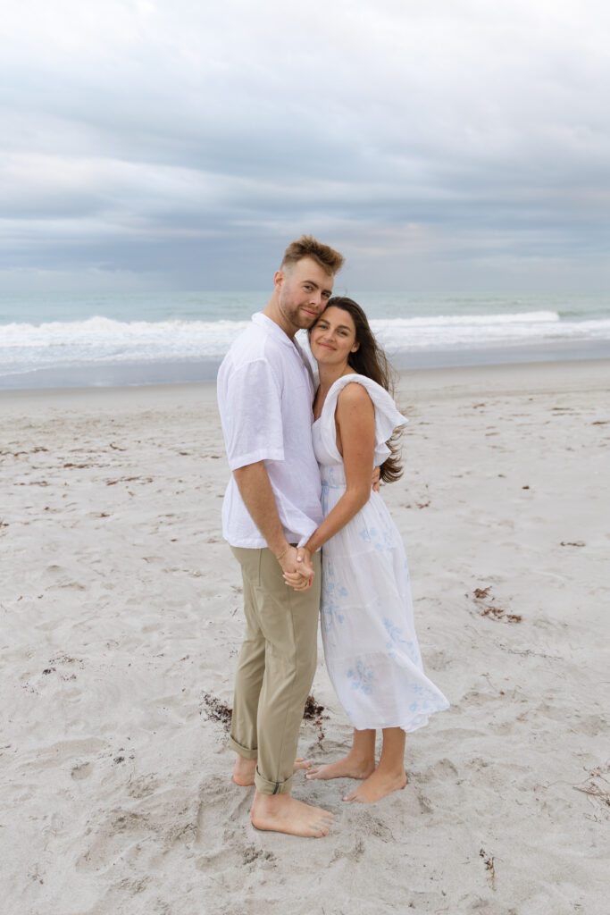 Engaged couple standing together on Cocoa Beach, with the ocean behind them, photographed by The Deans Photography.