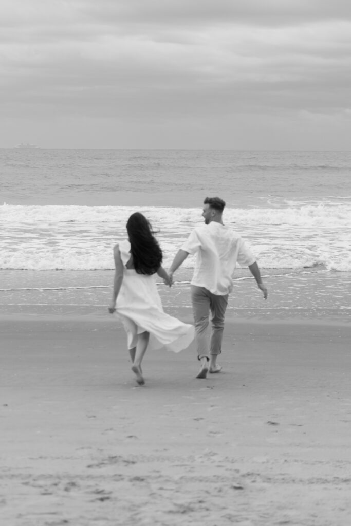 Engaged couple walking barefoot along Cocoa Beach at sunset, captured by The Deans Photo with the warm glow of the sun.