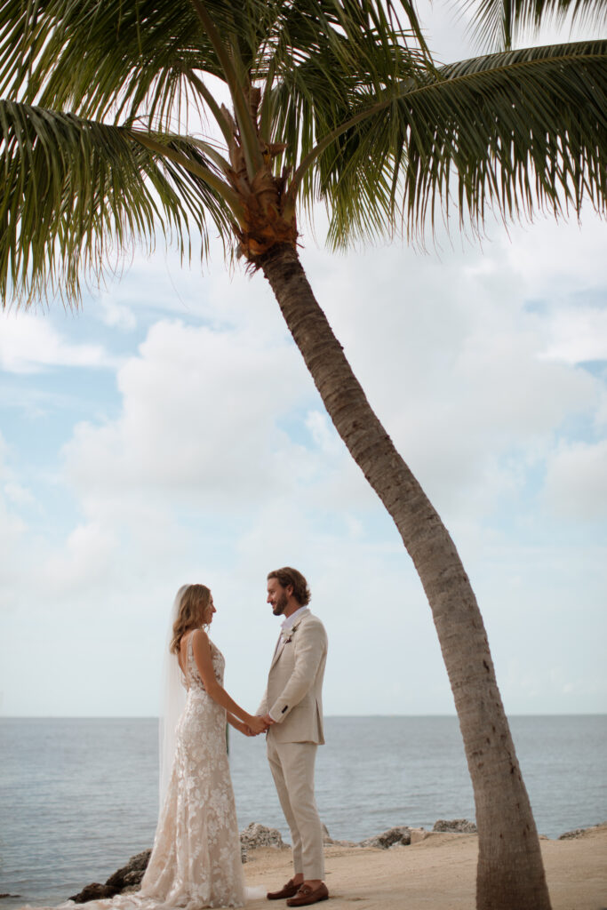 An intimate moment between the newly wedded couple holding hands in front of a tropical palm tress and the ocean photographed by The Deans Photography