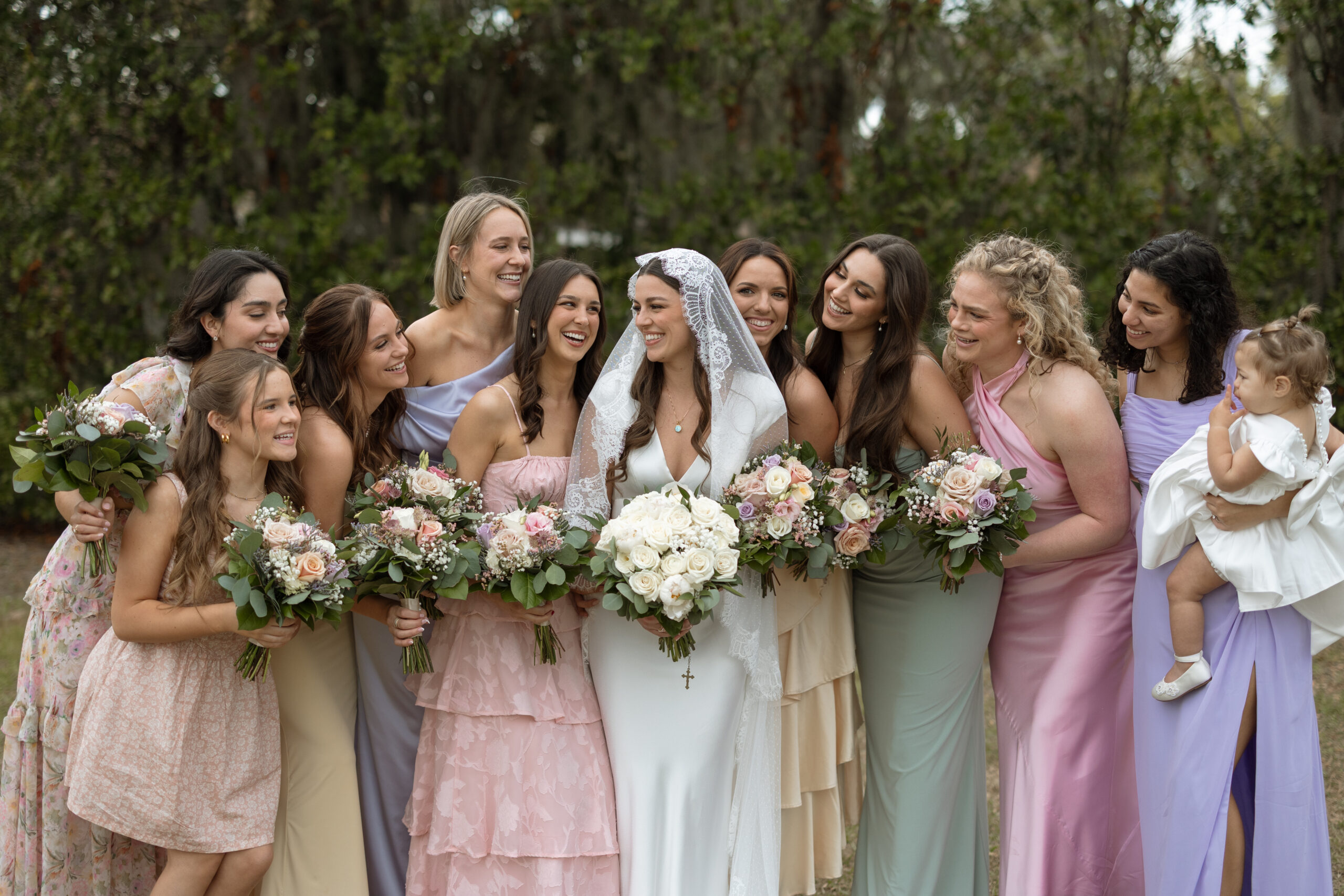 Bride and bridesmaids in elegant, flowing dresses pose in a lush Merritt Island garden, surrounded by soft florals and natural light
