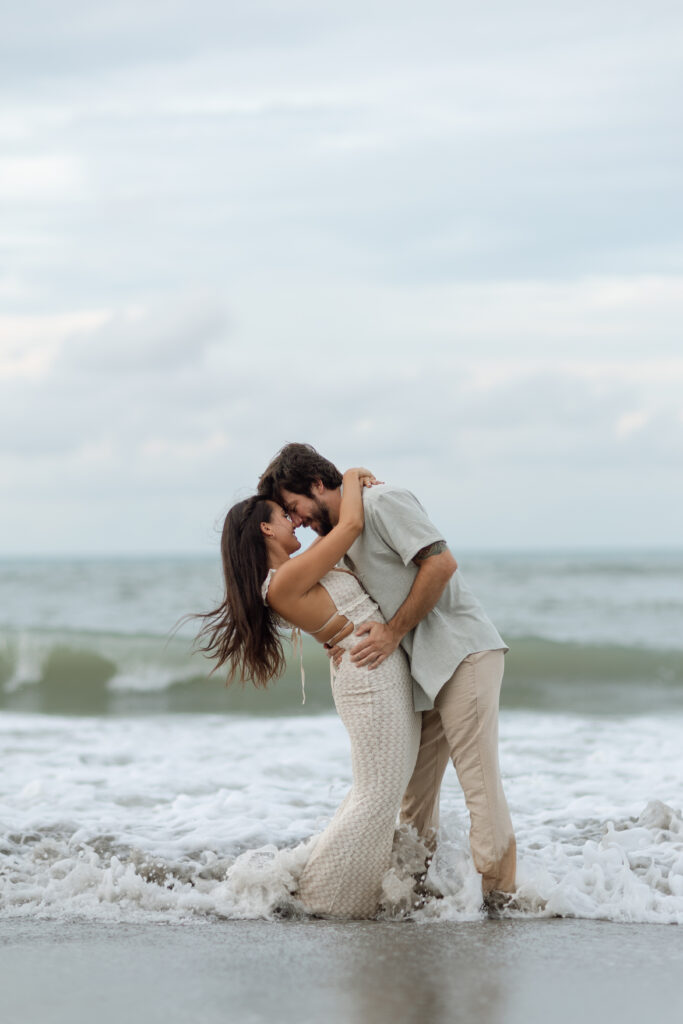 Couple walking barefoot along the shoreline at golden hour, wearing neutral-toned outfits that flow in the breeze.