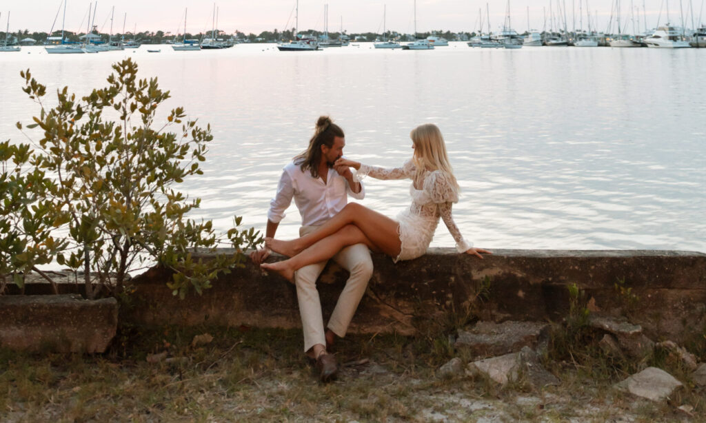 Couple holding hands, showing off engagement ring and textured linen suit with river in the background.