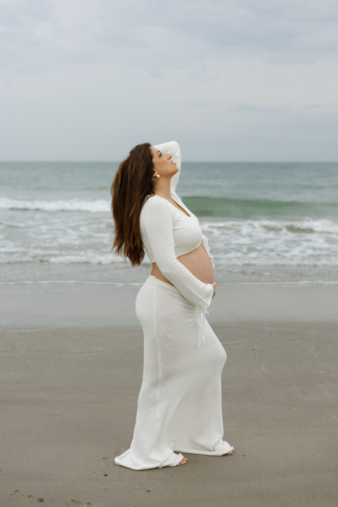 Pregnant woman in a flowy dress standing on Cocoa Beach at sunset during a maternity photo session