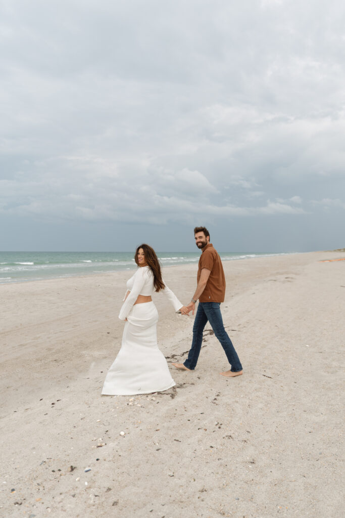Expecting couple smiling and holding hands during a Cocoa Beach maternity session at sunset