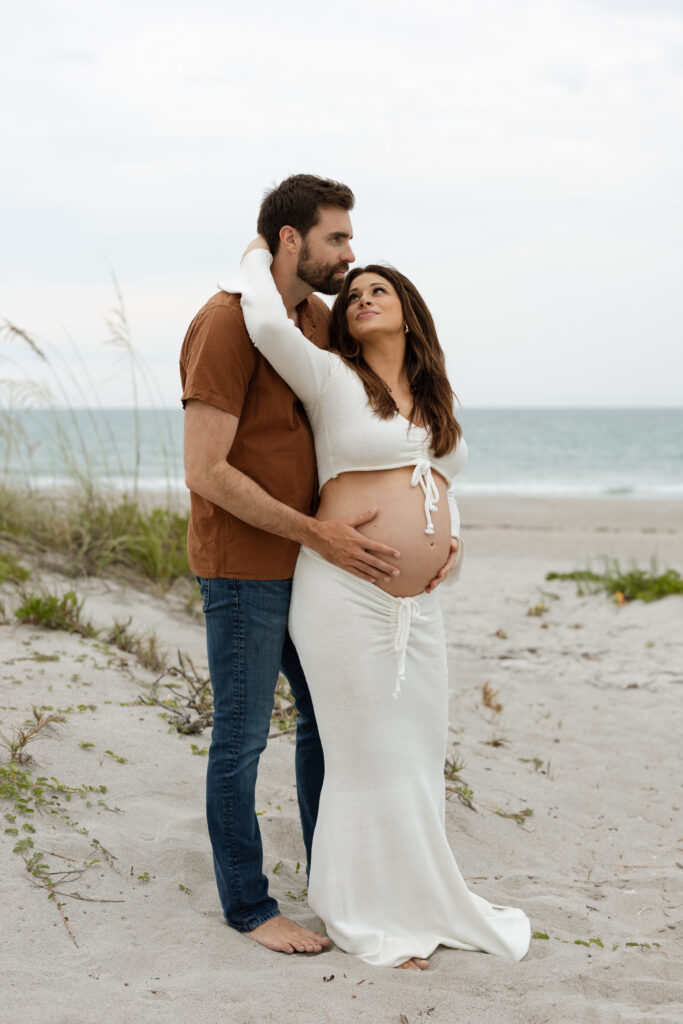 Expecting couple smiling and holding one another a Cocoa Beach maternity session at sunset